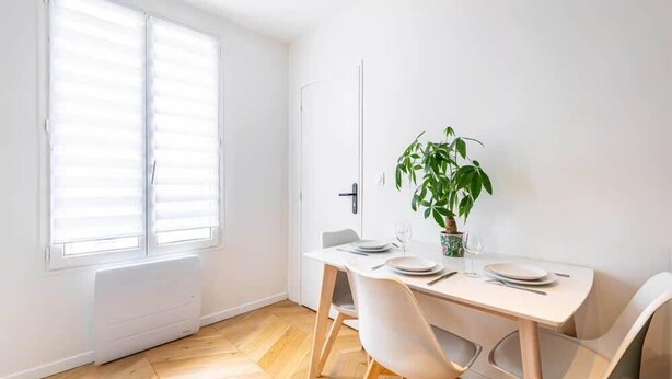 Un comedor con una mesa lista para dos comensales, una planta en maceta y un ventanal con persianas.