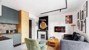 Un espace de vie moderne comprenant une cuisine, un coin repas et un coin salon avec un canapé et une table basse. Un téléviseur est fixé au mur, et un poster encadré y est également accroché.