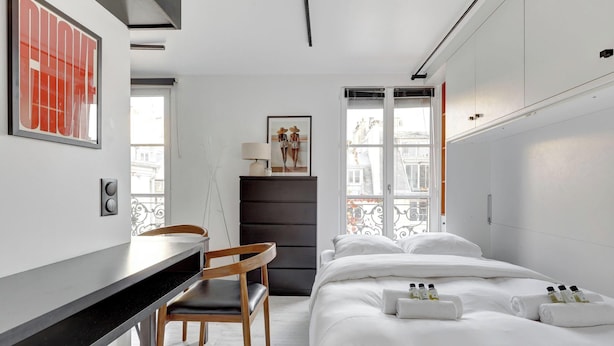 Une chambre d’hôtel comprenant un lit, un bureau, une chaise, une armoire et une fenêtre donnant sur des immeubles.