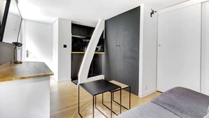 Ein kompakter, moderner Raum mit Schreibtisch aus Holz, einem kleinen schwarzen Tisch und einem grauen Sofa. Ein weißer Kleiderschrank und eine dunkelgraue Wand sind zu sehen.