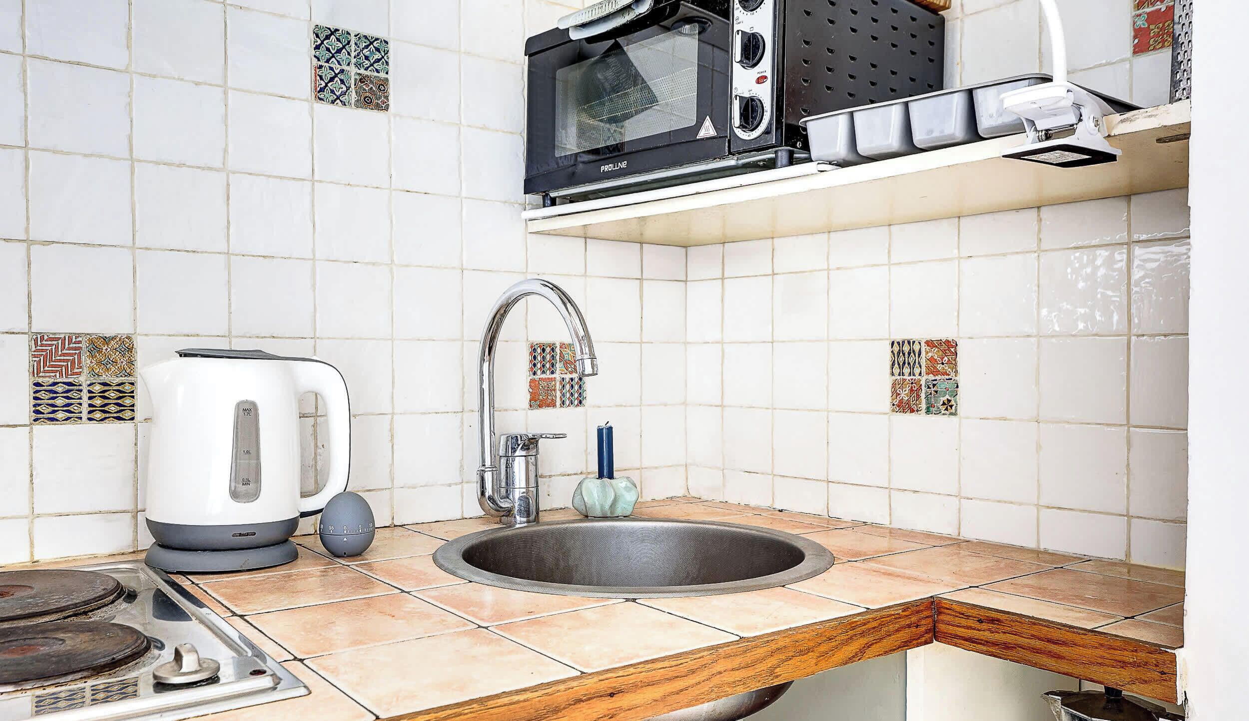 A kitchen with white tiles, a sink, a kettle, a stove, a microwave, and a faucet.