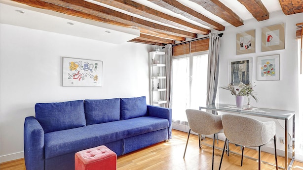 Un soggiorno con un divano blu, un poggiapiedi rosso, un tavolino da caffè in vetro e due poltrone. Si nota un soffitto in legno con travi a vista, una parete bianca con opere d'arte incorniciate e una finestra con tende.