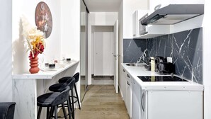 Una cocina estrecha con encimera blanca, salpicadero de mármol negro y estufa negra. Hay dos taburetes negros y un jarrón con flores secas a la izquierda.