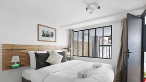 Una habitación de hotel moderna con un cabecero de madera, una cama grande con ropa de cama blanca, un cuadro enmarcado en la pared y una ventana con vistas al exterior.