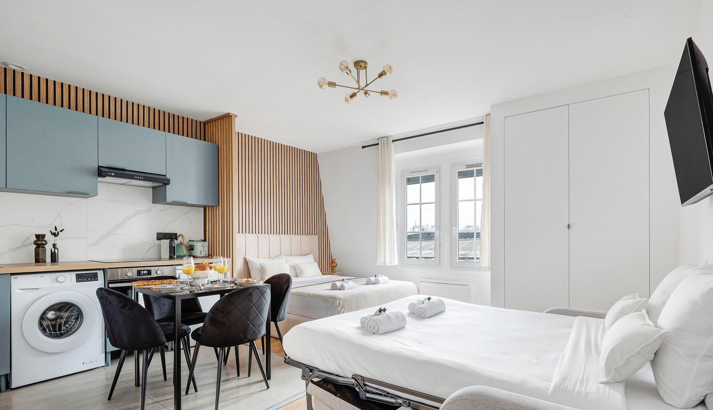 A modern hotel room with a bed, a dining area, a kitchen, and a washing machine.