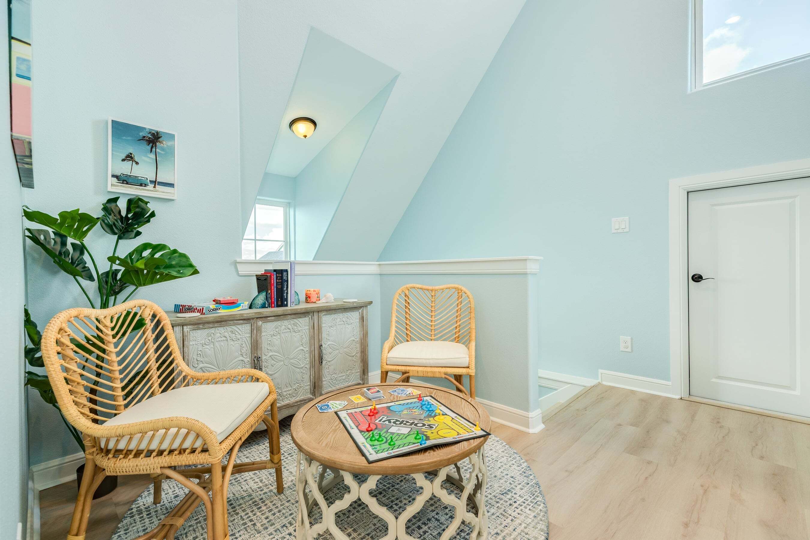 A room with a high ceiling, a rattan chair, a small table with a board game, a bookshelf, and a large plant.