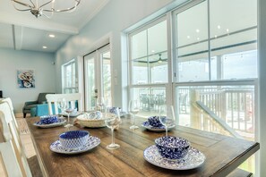 A dining area with a wooden table set for a meal, large windows, and a view of the outdoors. - Coastal Gem (Galveston)