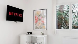 Moderni huone, jossa on seinälle asennettu televisio, jossa näkyy teksti ”NETFLIX”, kehystetty kukka-aiheinen taideteos ja ikkuna, josta avautuu näkymä puita kohti.