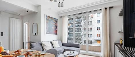 Una sala moderna con un sofá gris, una mesa de comedor con alimentos para el desayuno y grandes ventanales con vista a un edificio.