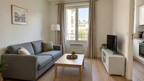 A modern living room with a grey sofa, a wooden coffee table, a television on a stand, and a view of buildings through a window. - Charming Paris Apartment · Balcony (Paris)