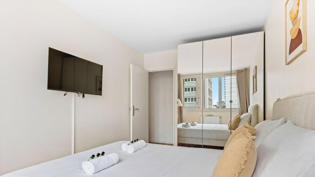 Ein Hotelzimmer mit Bett, einem an der Wand befestigten Fernseher, einem großen Spiegel und einem Fenster mit Blick auf Gebäude.