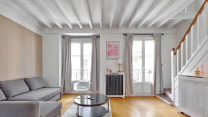 A modern living room with a grey sofa, a round coffee table, and a radiator. There are two windows with curtains, a staircase, and a framed artwork on the wall. - Magnifique duplex avec terrasse et vue imprenable sur la Tour Eiffel (Paris)