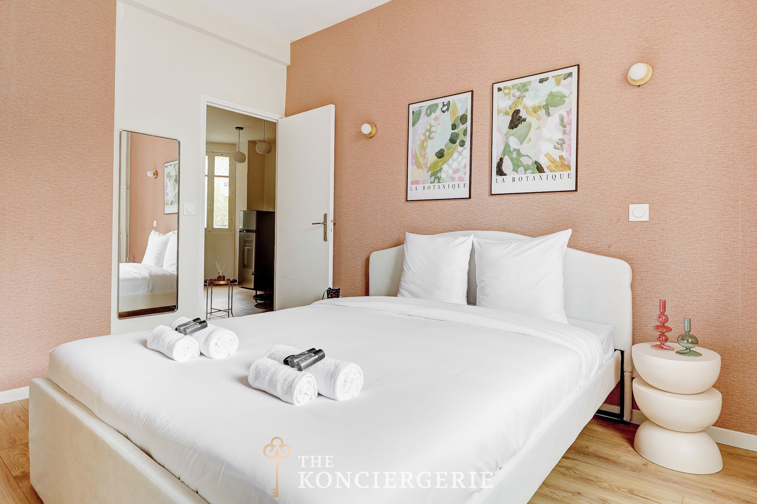 A hotel room with a large bed, white linens, a bedside table with a vase, and two framed botanical posters on the wall.