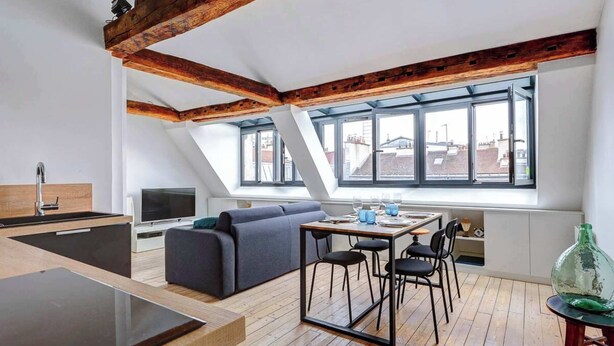 En moderne loftlejlighed med synlige træbjælker, et spiseområde med bord og stole, en sofa og et stort vindue med udsigt over byen.