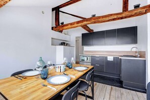 Ein Essbereich mit einem Holztisch, gedeckt für vier Personen, eine Küche mit dunklen Schränken und freiliegenden Holzbalken.