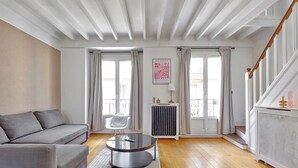 Ein modernes Wohnzimmer mit grauem Sofa, einem runden Couchtisch und einer Heizung. Zwei Fenster mit Vorhängen, eine Treppe und ein gerahmtes Kunstwerk sind an der Wand zu sehen.