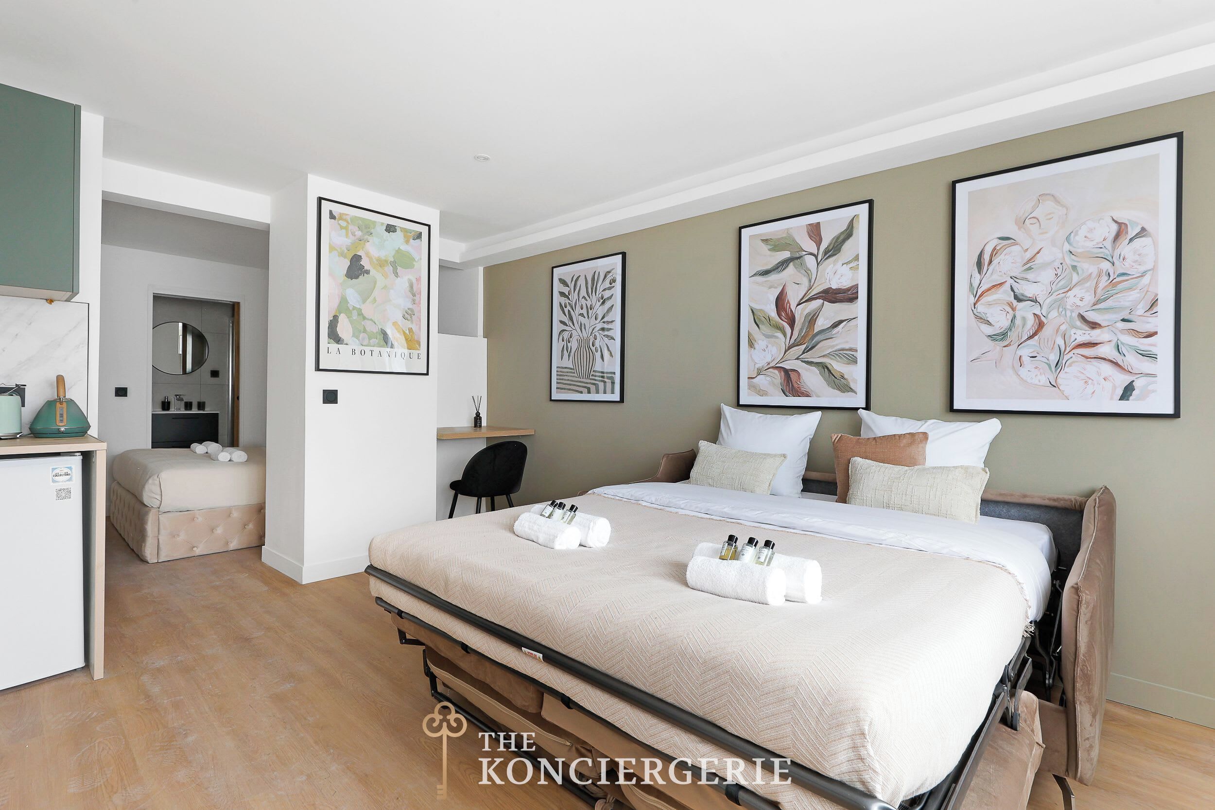 Habitación de hotel moderna con una cama grande, un escritorio y una pequeña kitchenette. Hay obras de arte enmarcadas en las paredes y un minibar en la esquina.