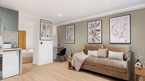 Una sala de estar moderna con un sofá color beige, un escritorio pequeño y una kitchenette. Hay tres láminas botánicas enmarcadas en la pared.