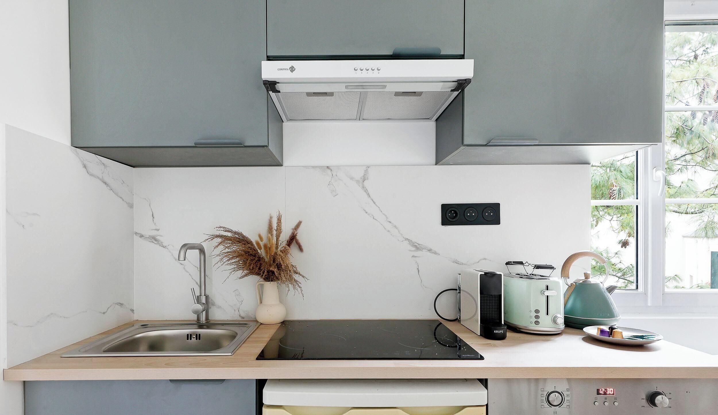 Une cuisine moderne avec des meubles gris, un crédence blanche et une fenêtre donnant sur les arbres.