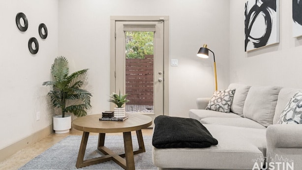 A modern living room with a round wooden coffee table, a grey sofa with patterned cushions, a black throw blanket, a floor lamp, and a potted plant.