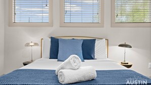 A neatly made bed with a blue and white patterned pillow, a white towel, and a blue textured blanket. There are two bedside lamps on wooden nightstands. Three windows with blinds are visible, allowing natural light into the room.