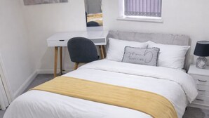 Een slaapkamer met een bed, een bureau, een stoel, een nachtkastje, een lamp, een raam met gordijnen en een spiegel aan de muur.