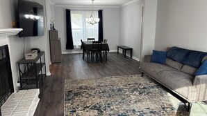 Un salon doté d’un parquet en bois foncé, d’un canapé gris avec des coussins bleus, d’un tapis à motifs, d’un coin repas avec une table et des chaises, et d’un téléviseur fixé au mur.