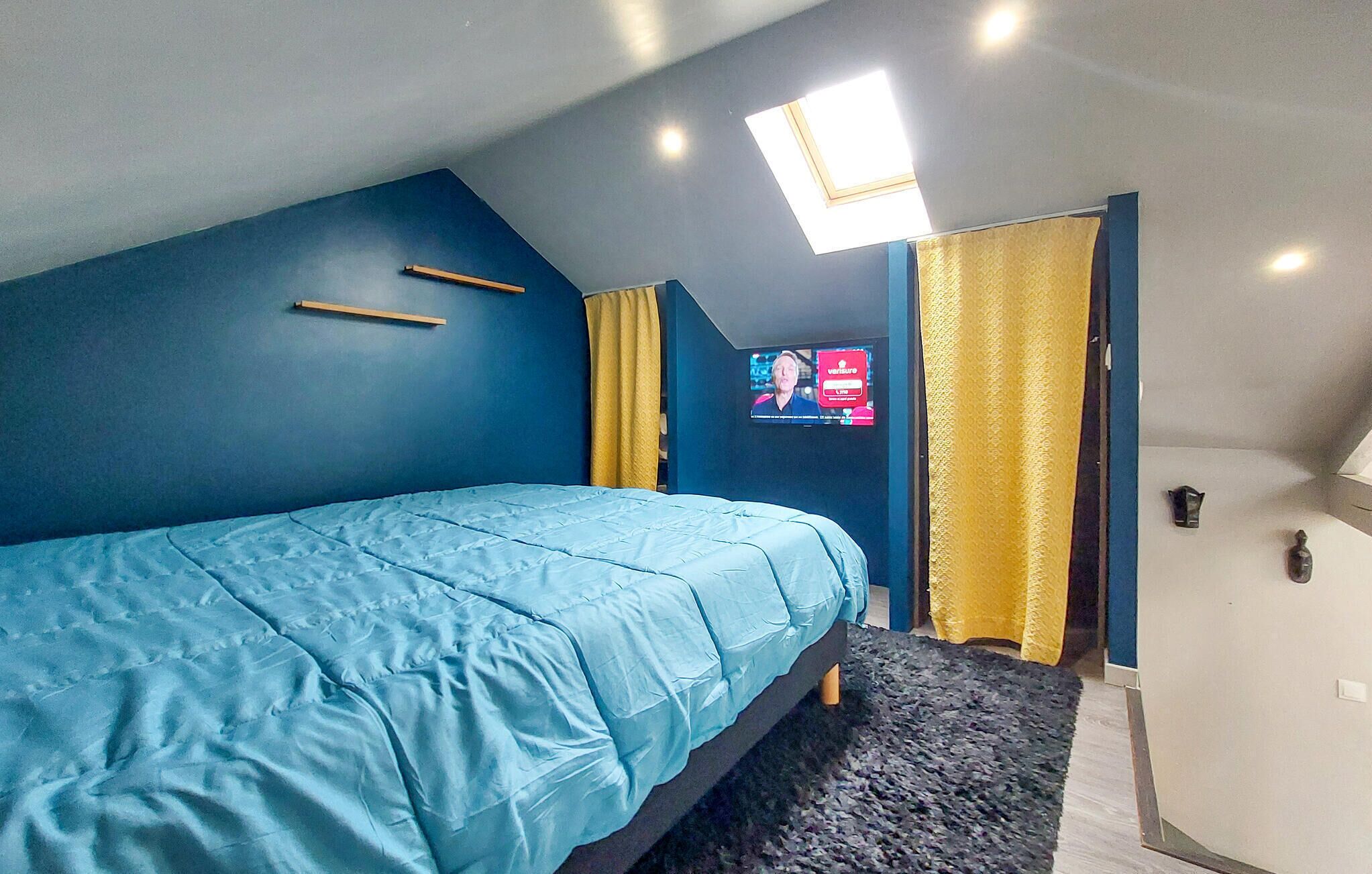 Un dormitorio con cama, televisor, tragaluz y puerta con cortinas.