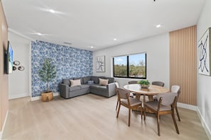 Een moderne woonkamer met een grijze hoekbank, een ronde houten eettafel met stoelen, een groot raam met uitzicht en een behang met patroon.