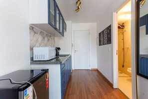 A narrow hallway with a kitchen area on the left, featuring a microwave and cabinets. A door leads to a bathroom with a shower and toilet visible. - Studio 402 Perdizes 900m Allianz Parque c Academia (São Paulo)