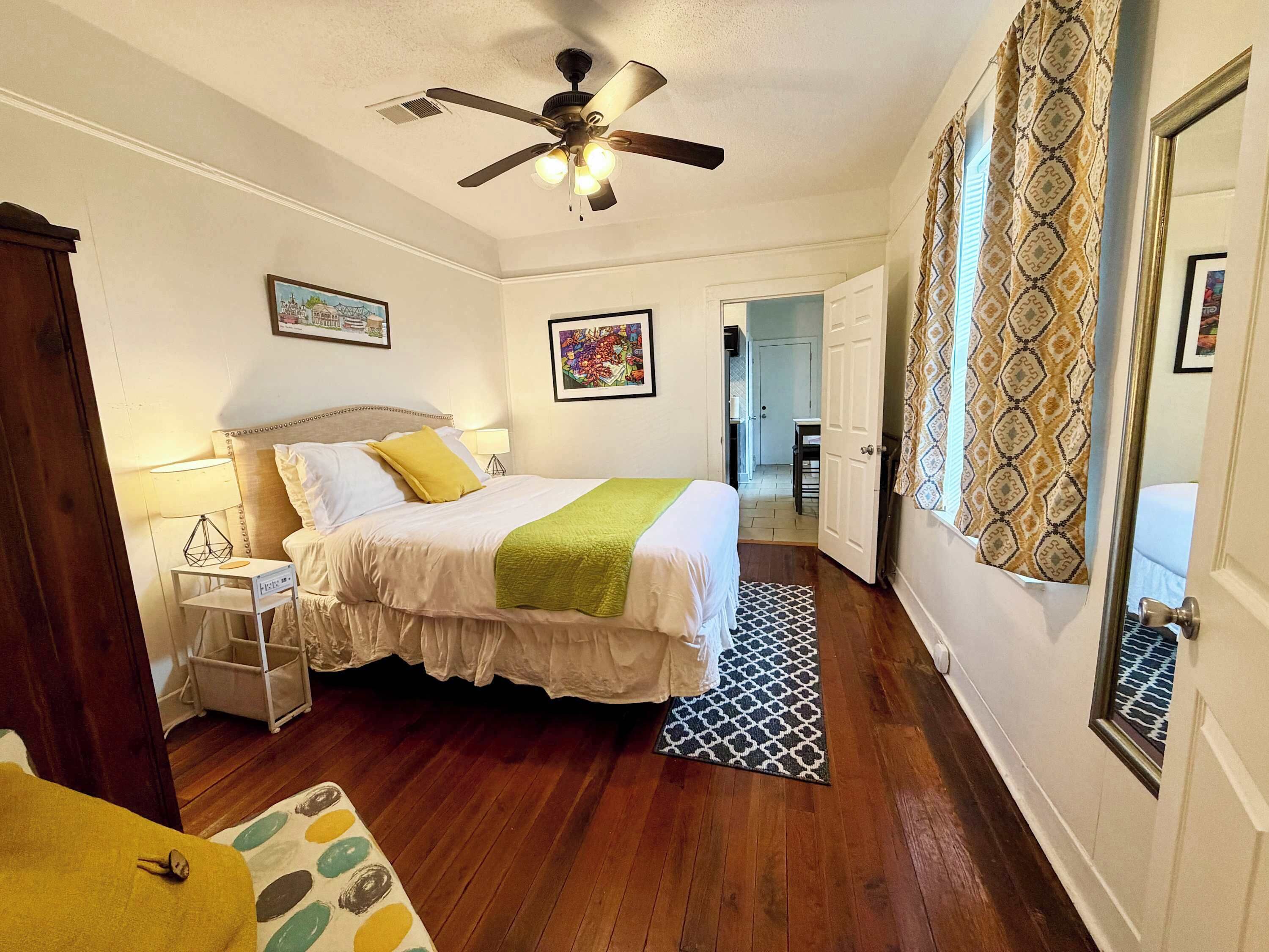 Una habitación de hotel con cama, ventilador de techo, mesita de noche, alfombra estampada y una puerta que da a otra habitación.