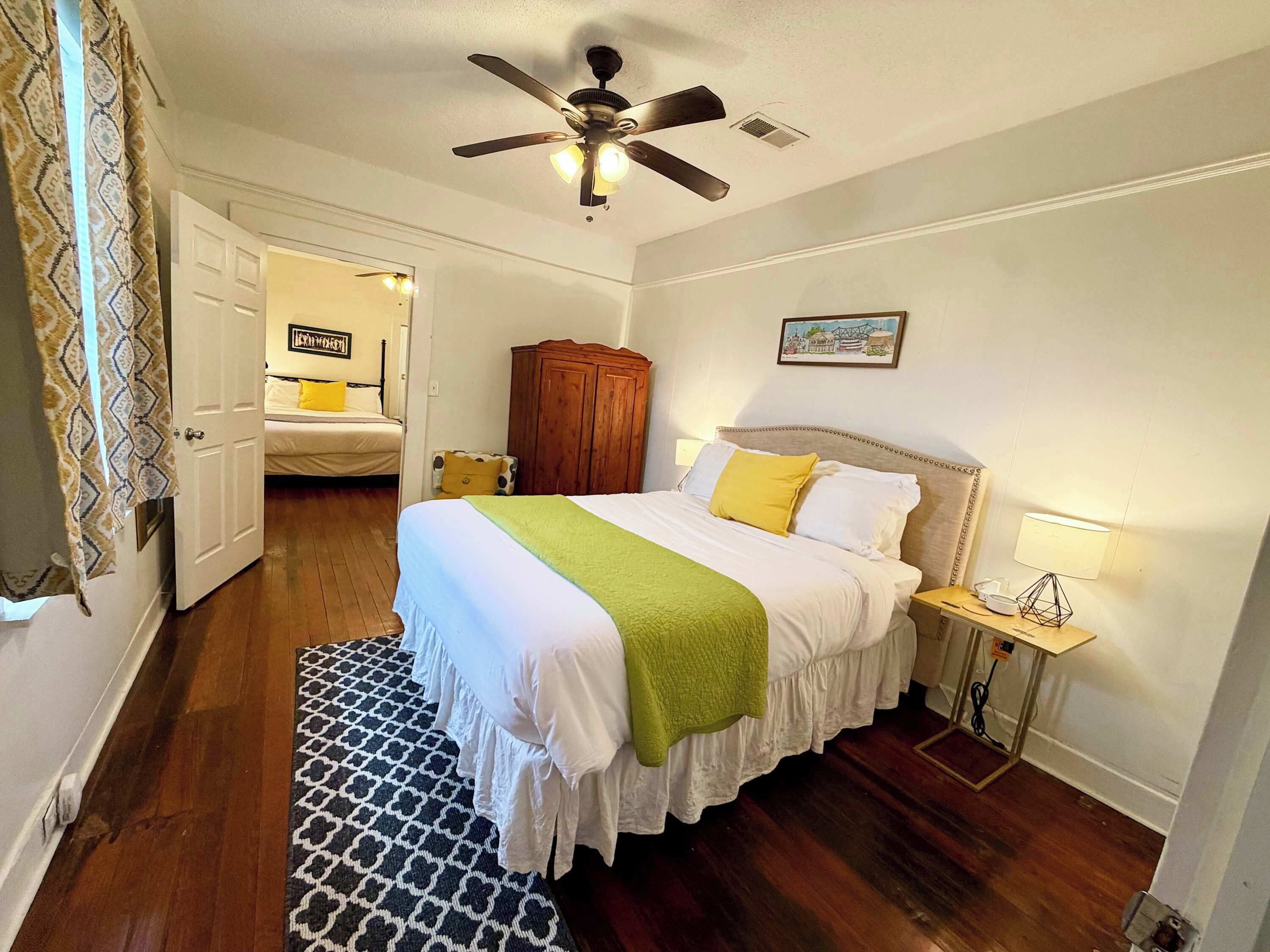 Une chambre à coucher comprenant un lit, un ventilateur de plafond, une armoire en bois, une table de chevet avec une lampe et un tapis à motifs.