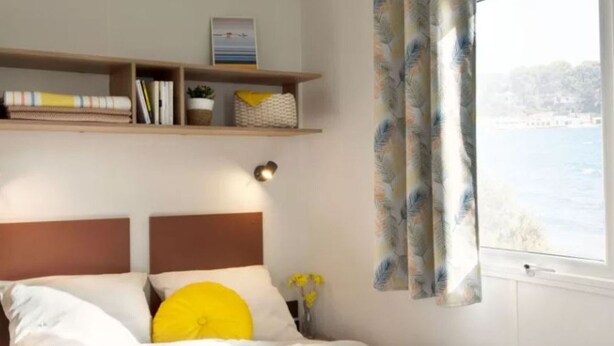A bedroom with a bed, a nightstand, a shelf with books and decorative items, and a window with curtains. - Spacious mobile home - 8 guests (Visan)