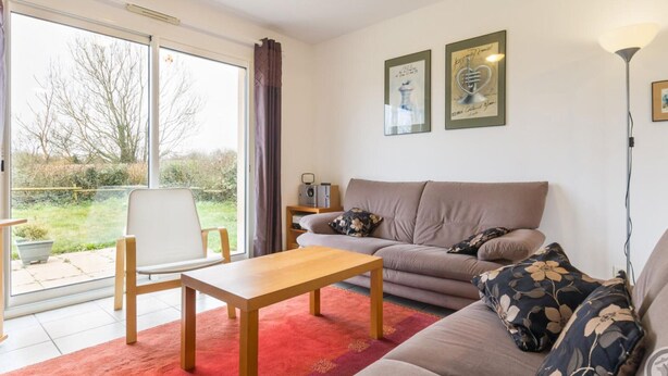 Ein Wohnzimmer mit einer Couch, einem Holztisch und einem roten Teppich. Ein Fenster bietet Blick ins Freie.