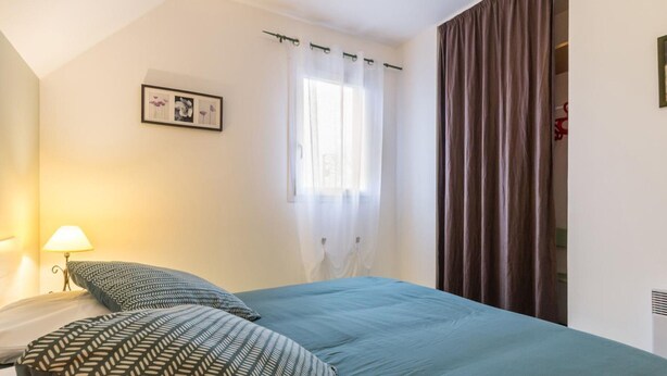 Quarto com cama, mesa de cabeceira, luminária, janela com cortinas e quadro pendurado na parede.