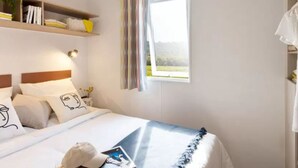 Een slaapkamer met een bed, een nachtkastje, een plank met boeken en een plant, een raam met gordijnen en uitzicht op de omgeving.