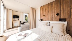 Ein modernes Schlafzimmer mit einem großen Bett, einem hölzernen Kopfteil, einem an der Wand befestigten Fernseher und einem Essbereich, der durch eine offene Tür sichtbar ist.