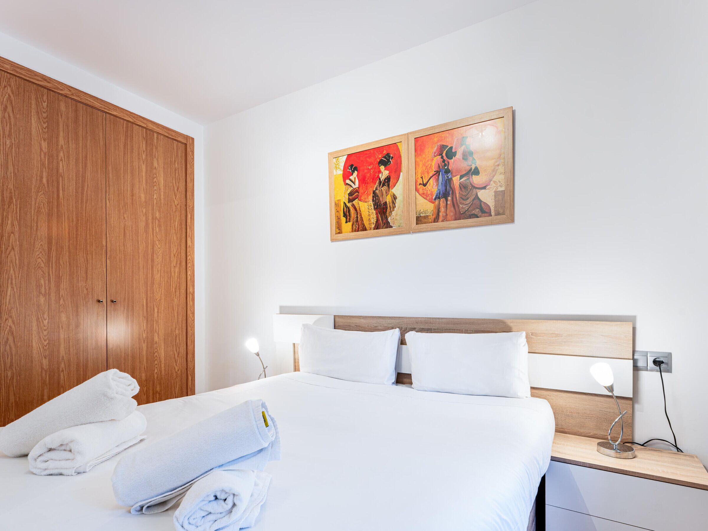 A hotel room with a bed, two paintings on the wall, a wooden wardrobe, and a nightstand with a lamp.