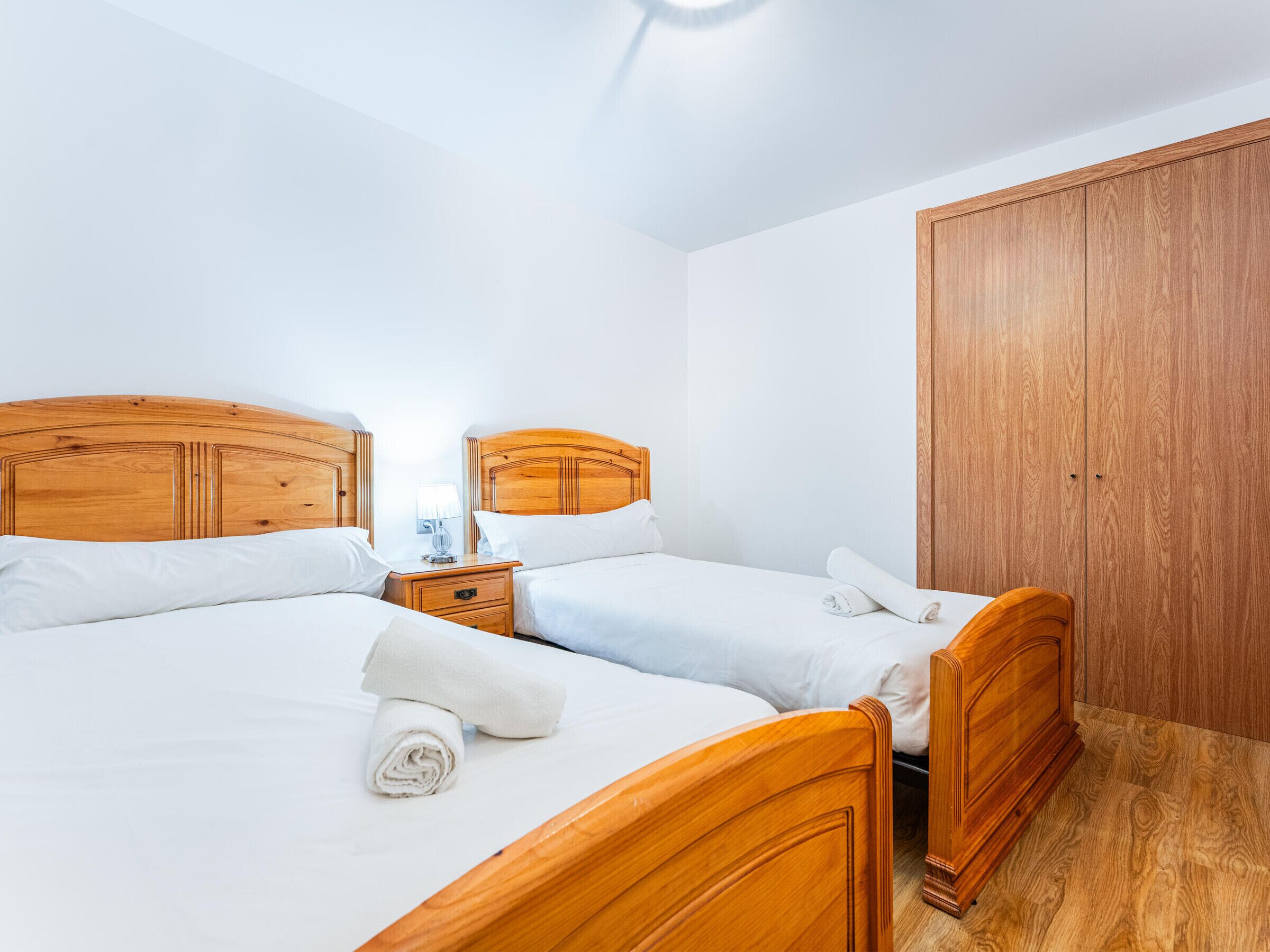 A hotel room with two wooden beds, a nightstand with a lamp, and a wooden wardrobe.