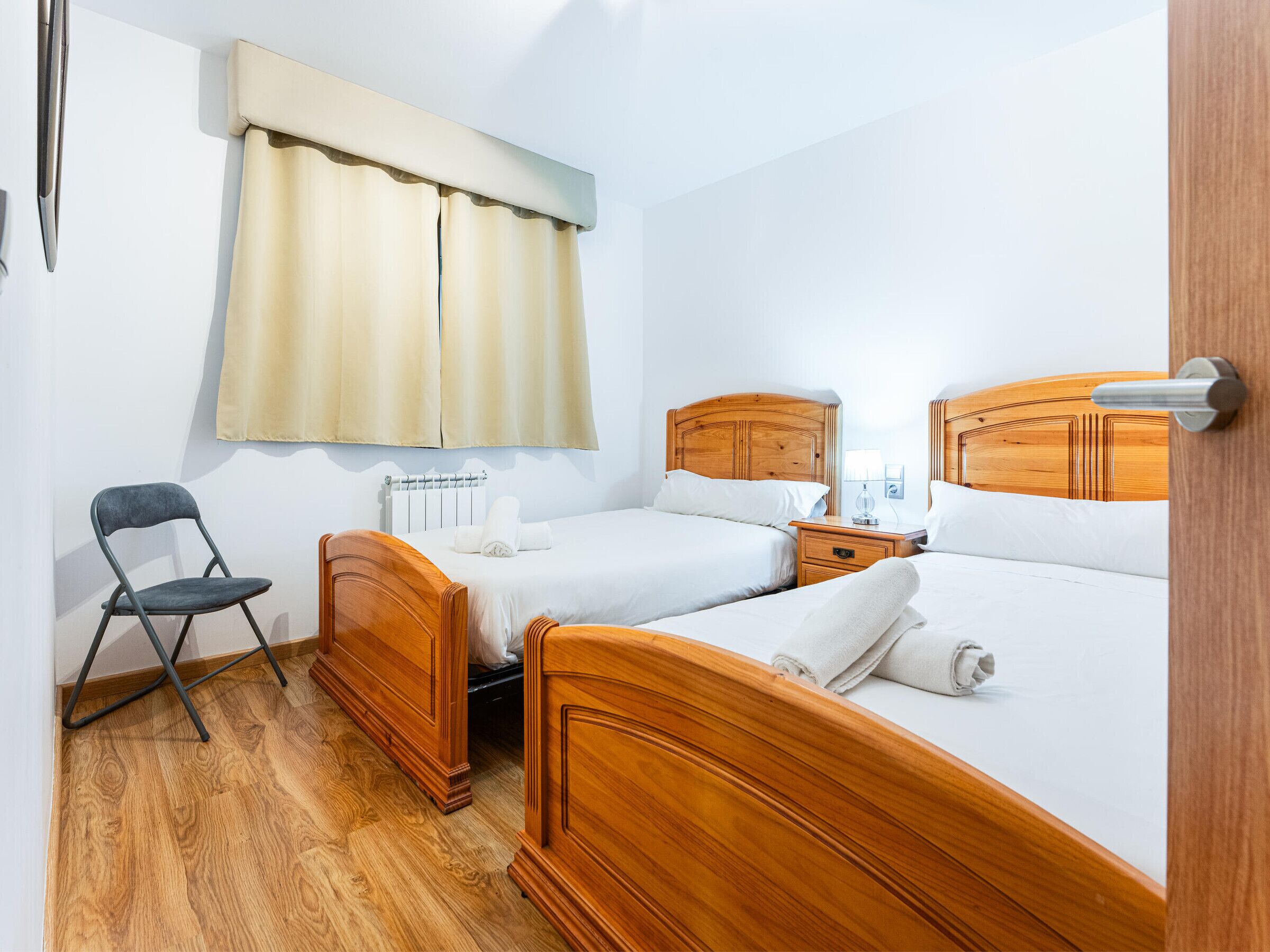 Ein Hotelzimmer mit zwei Holzbett, einem Sessel, einem Nachttisch und einem Fenster mit Vorhängen.