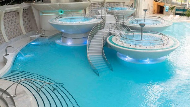 Una piscina interior con varios jacuzzis, una escalera central y un techo de cristal.