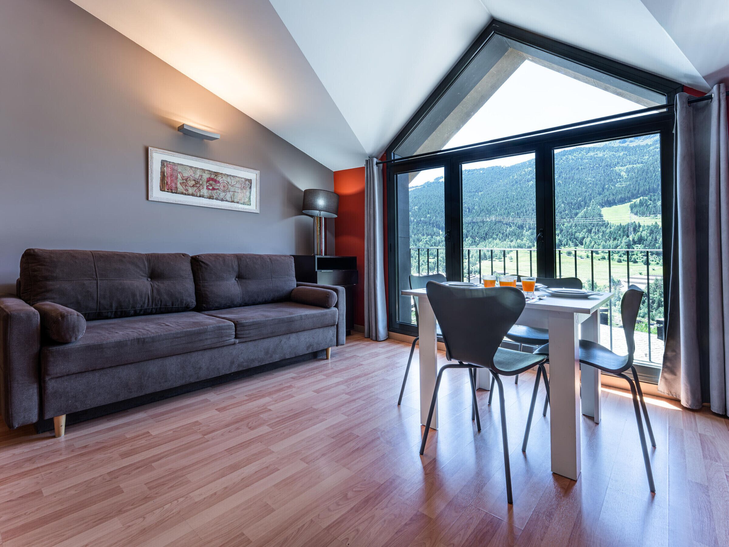 Un salon moderne avec un canapé gris, une table à manger avec des chaises, une lampe sur pied et une grande fenêtre donnant sur un paysage montagneux.