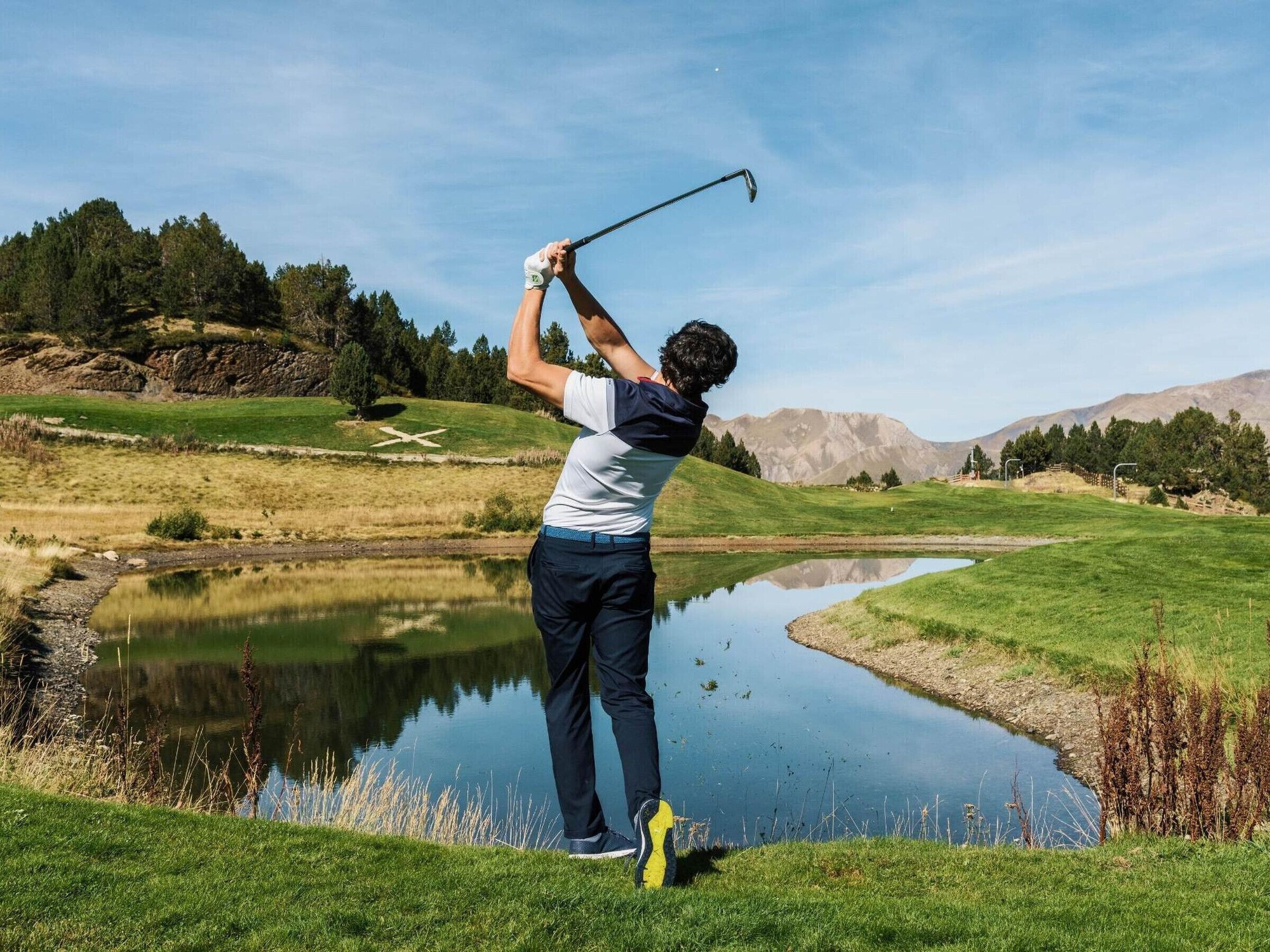 A golf course with a pond, green hills, and a clear sky.