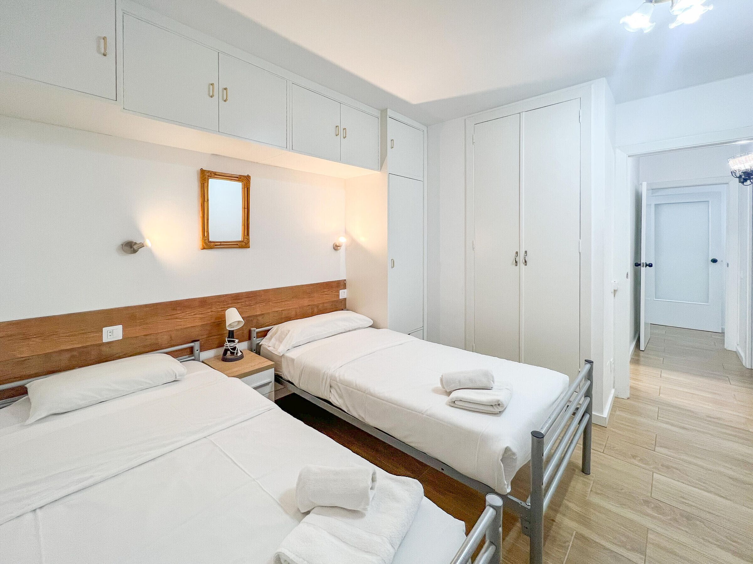 A hotel room with two single beds, a wooden headboard, a nightstand with a lamp, and a wardrobe with a mirror.