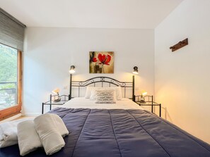 Ein ordentlich bezogenes Bett mit dunkelblauer Decke und weißen Kissen, zwei Nachttischlampen, ein Gemälde mit roten Blumen an der Wand und ein Fenster mit Blick auf Grünflächen.
