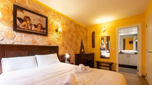 Habitación de hotel con pared de piedra, cama con ropa blanca, mesita de noche con lámpara, cómoda y baño visible a través de una puerta abierta.