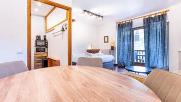 Habitación de hotel con una mesa de comedor de madera, una cama con ropa blanca, una pequeña kitchenette y vistas al exterior a través de una ventana con cortinas.