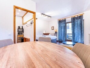 Habitació d'hotel amb taula de dinar de fusta, llit amb roba de llit blanca, petita cuineta i vista a l'exterior a través d'una finestra amb cortines.
