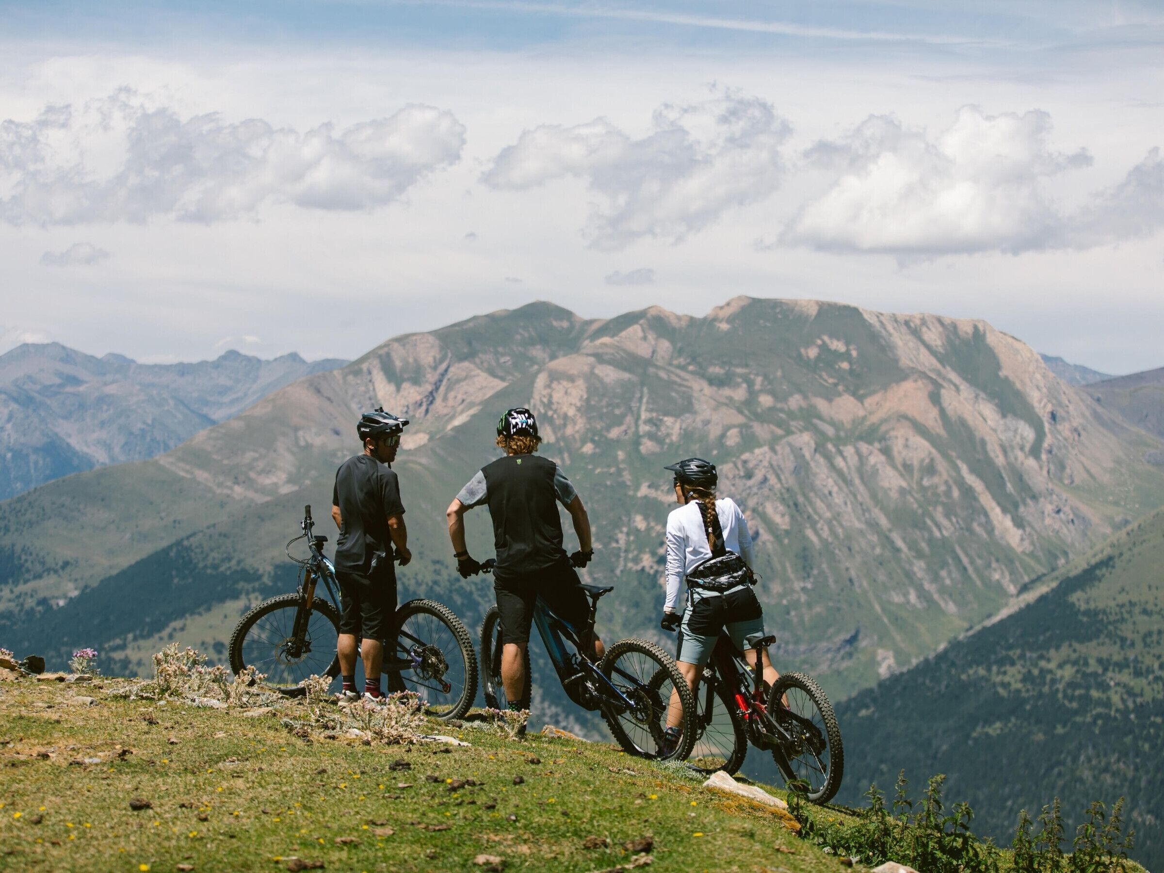 Tres personas en una colina cubierta de hierba con bicicletas, contemplando un paisaje montañoso.