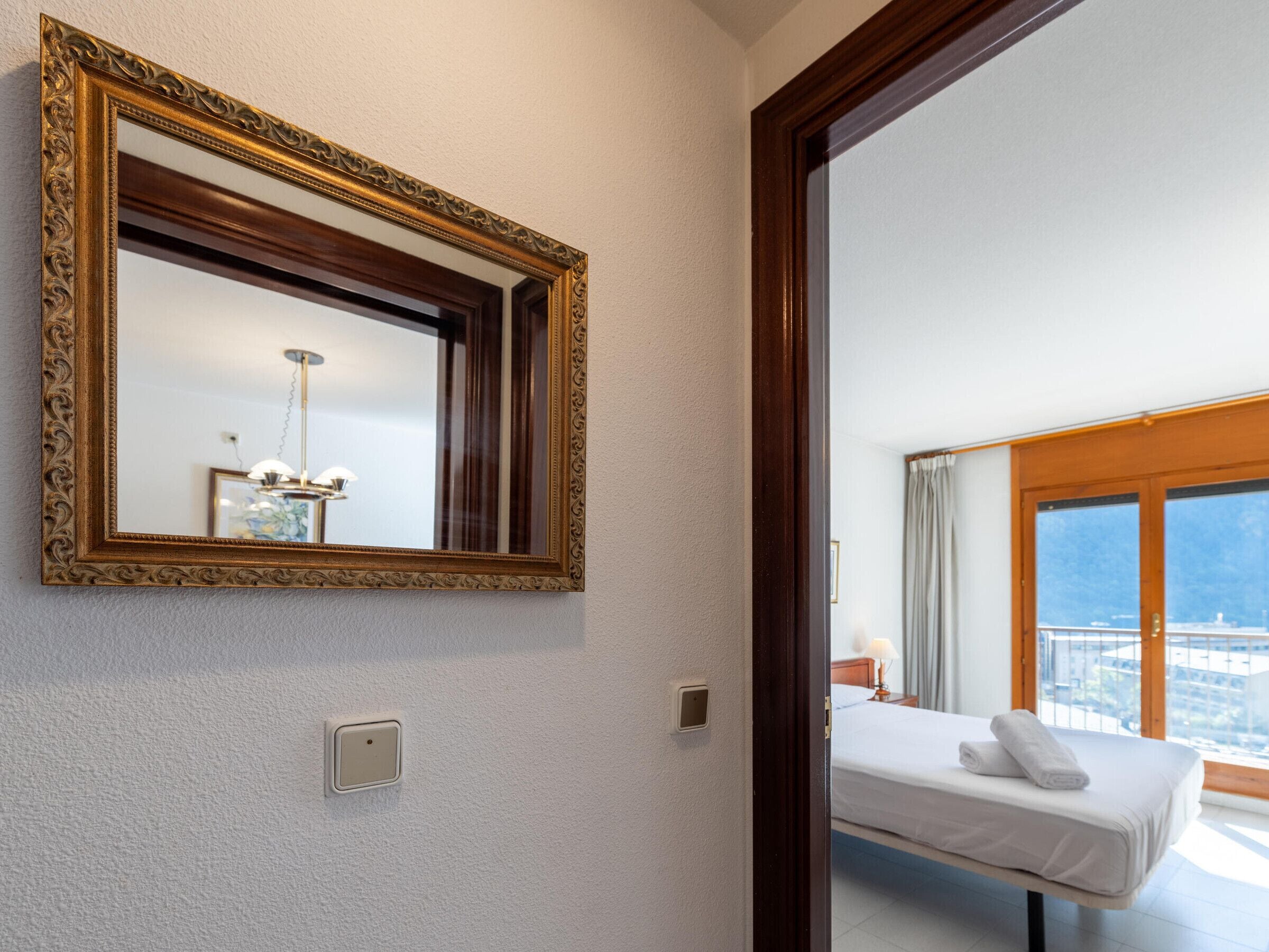 Hotelkamer met een grote, versierde spiegel, een bed met witte lakens, een nachtkastje met een lamp en uitzicht op de stad door grote ramen.